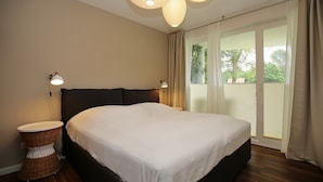 A bedroom with a large bed, two bedside lamps, a wicker side table, and a view of greenery through the window. - Likedeeler Wohnung 06 (Ostseebad Boltenhagen)