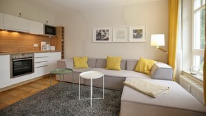 En moderne stue med sofa, rundt bord og et tekøkken med mikroovn og ovn.