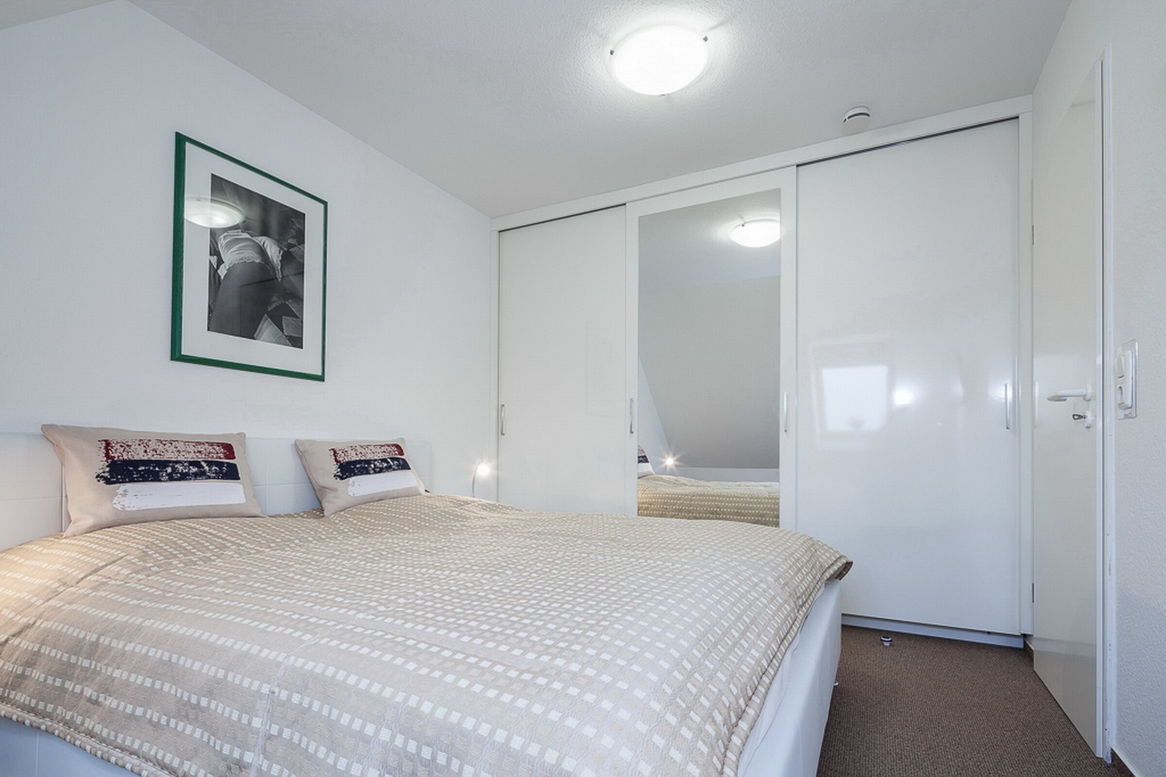 A bedroom with a bed, pillows, a framed picture, a wardrobe, and a ceiling light.