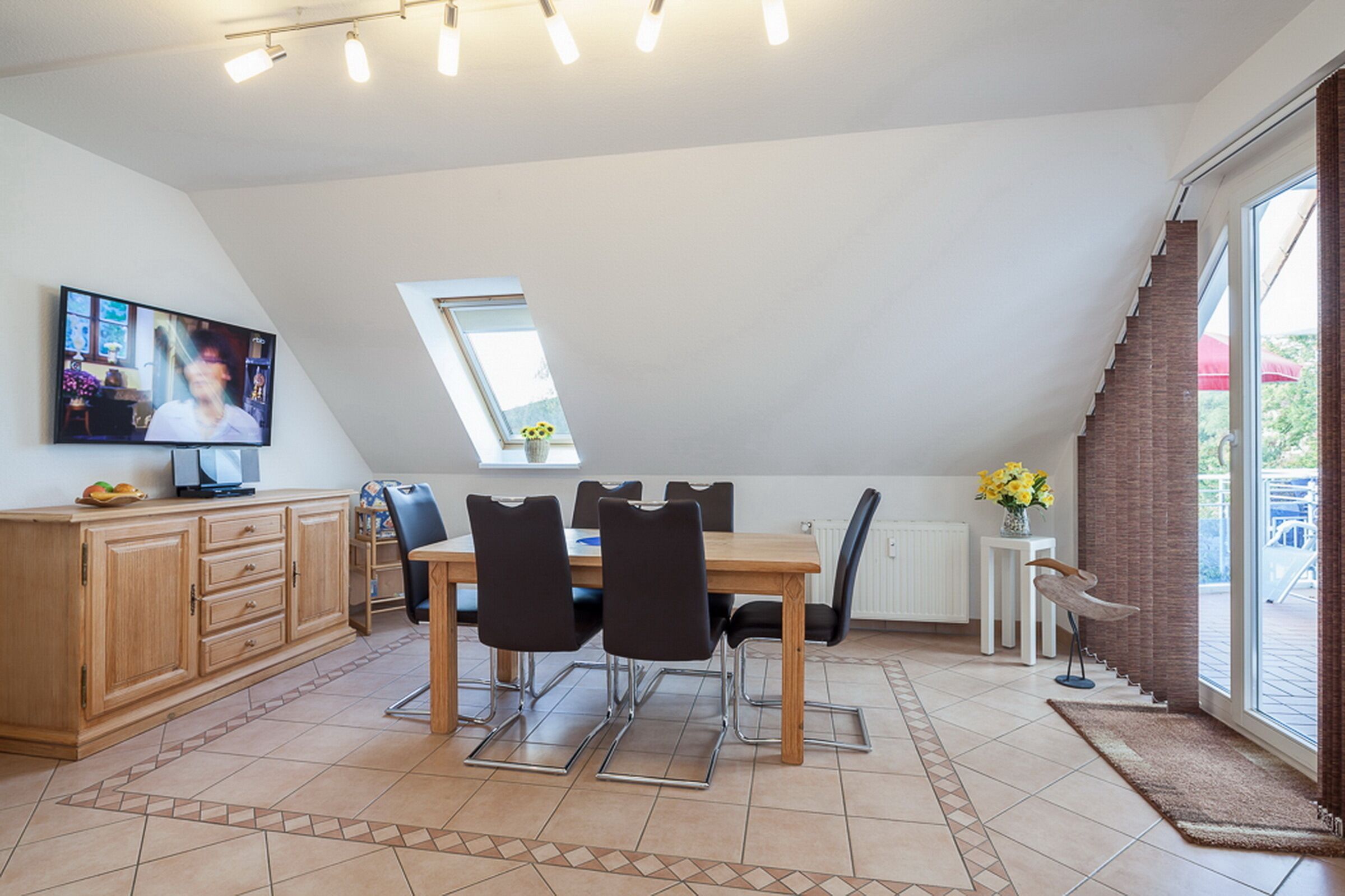 A dining area with a wooden table and chairs, a television on the wall, and a window with a view of the outdoors.