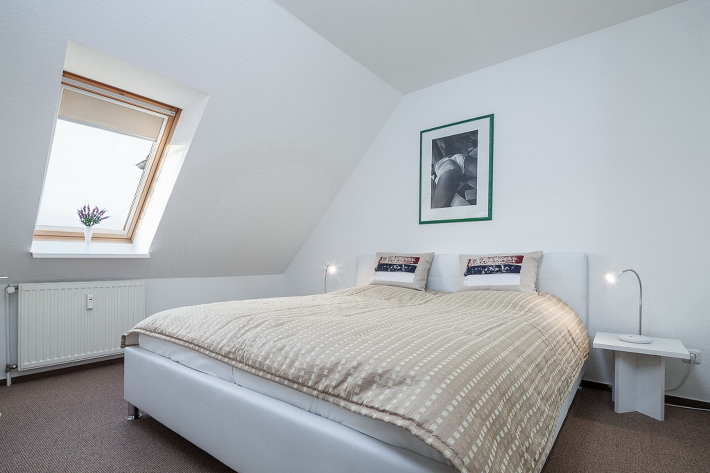 Une chambre à coucher avec un lit, une fenêtre, un tableau au mur et une table de chevet avec une lampe.