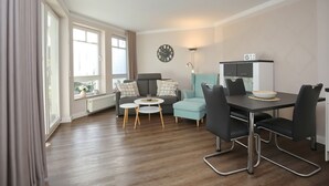 A modern living room with a dining area, large windows, a clock, and a radiator. - Likedeeler Wohnung 08 (Ostseebad Boltenhagen)