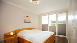 A bedroom with a wooden bed, a nightstand with a lamp, a window with a view of greenery, and a ceiling light fixture. - Likedeeler Wohnung 08 (Ostseebad Boltenhagen)