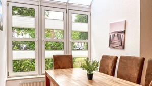 A dining area with a wooden table, six chairs, a potted plant, and a view of greenery through large windows. - Sünnslag Wohnung 131 (Ostseebad Boltenhagen)