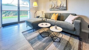 Una sala de estar moderna con un sofá seccional gris, dos mesitas auxiliares, un cuadro de veleros y un gran ventanal corredero que da a un jardín.
