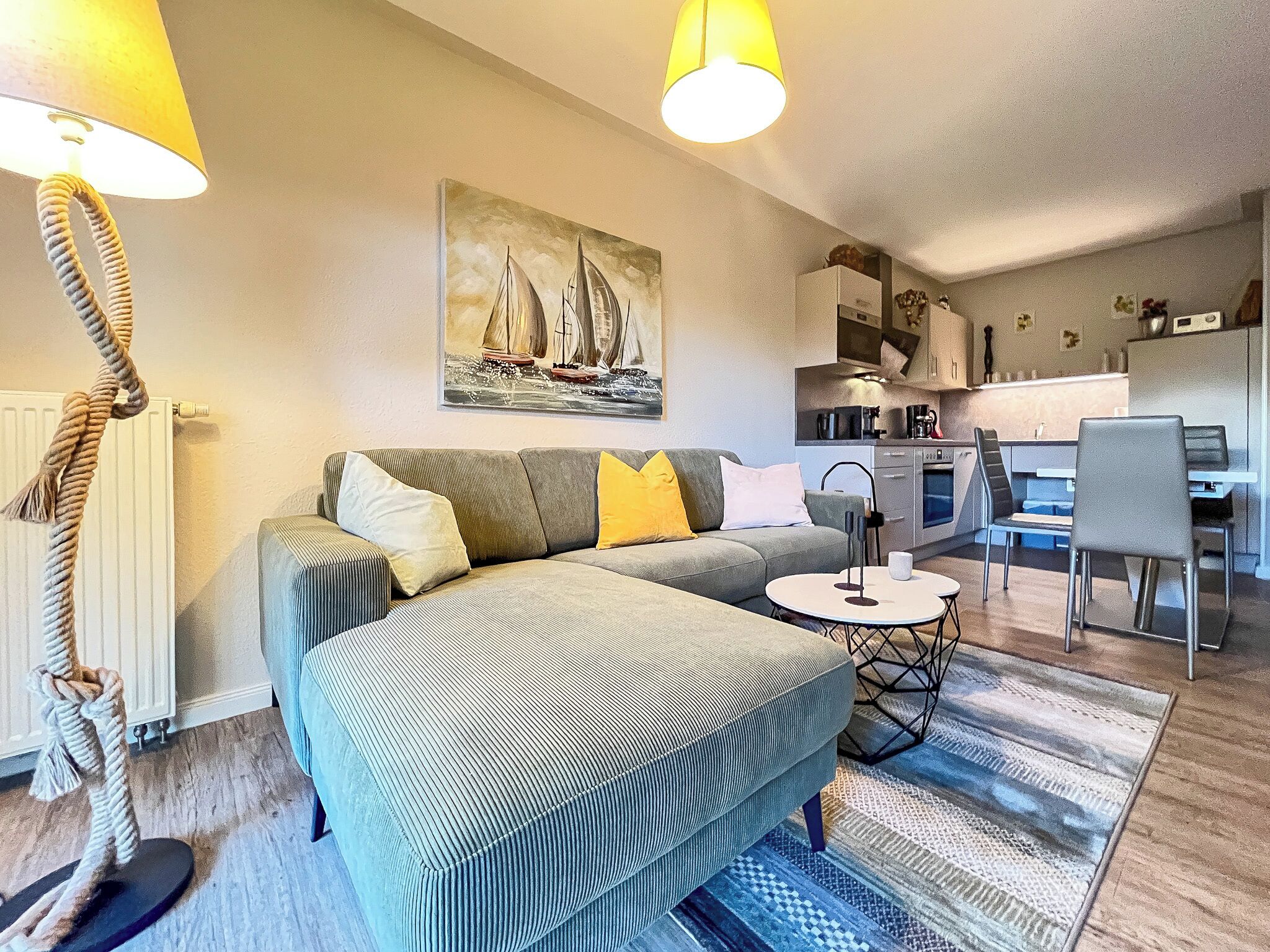 A modern living room with a grey sofa, a coffee table, and a dining area with a chair and table. There is a painting of sailboats on the wall, a lamp, and a kitchen in the background.