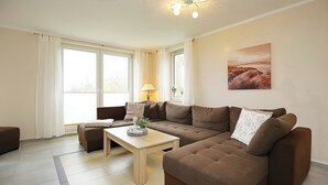Ein modernes Wohnzimmer mit einem braunen Ecksofa, einem Holztisch und einer Lampe. Große Fenster mit weißen Vorhängen, ein Bild an der Wand und eine Deckenleuchte sind ebenfalls vorhanden.