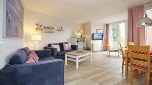 A living room with a sofa, coffee table, and wooden chairs. There is a TV, a lamp, and a shelf with decorative items. - Sünnslag Wohnung 122 (Ostseebad Boltenhagen)