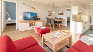 A modern living room with red sofas, a wooden coffee table, and a TV. There is a dining area with a table and chairs, and a kitchen with white cabinets and a sink. - Sünnslag Wohnung 127 (Ostseebad Boltenhagen)