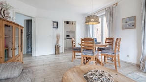 En rummelig opholdsstue med et træbord, stole og en sofa. Et køkken ses i baggrunden, og der er et stort vindue med gardiner.