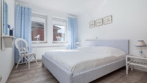 Et soveværelse med en stor seng, en stol, et vindue med gardiner og en væg med billeder i rammer.