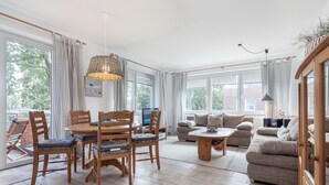 En rummelig opholdsstue med spisebord, stole, sofa og sofabord. Store vinduer med gardiner, en pendel og udsigt til det fri.
