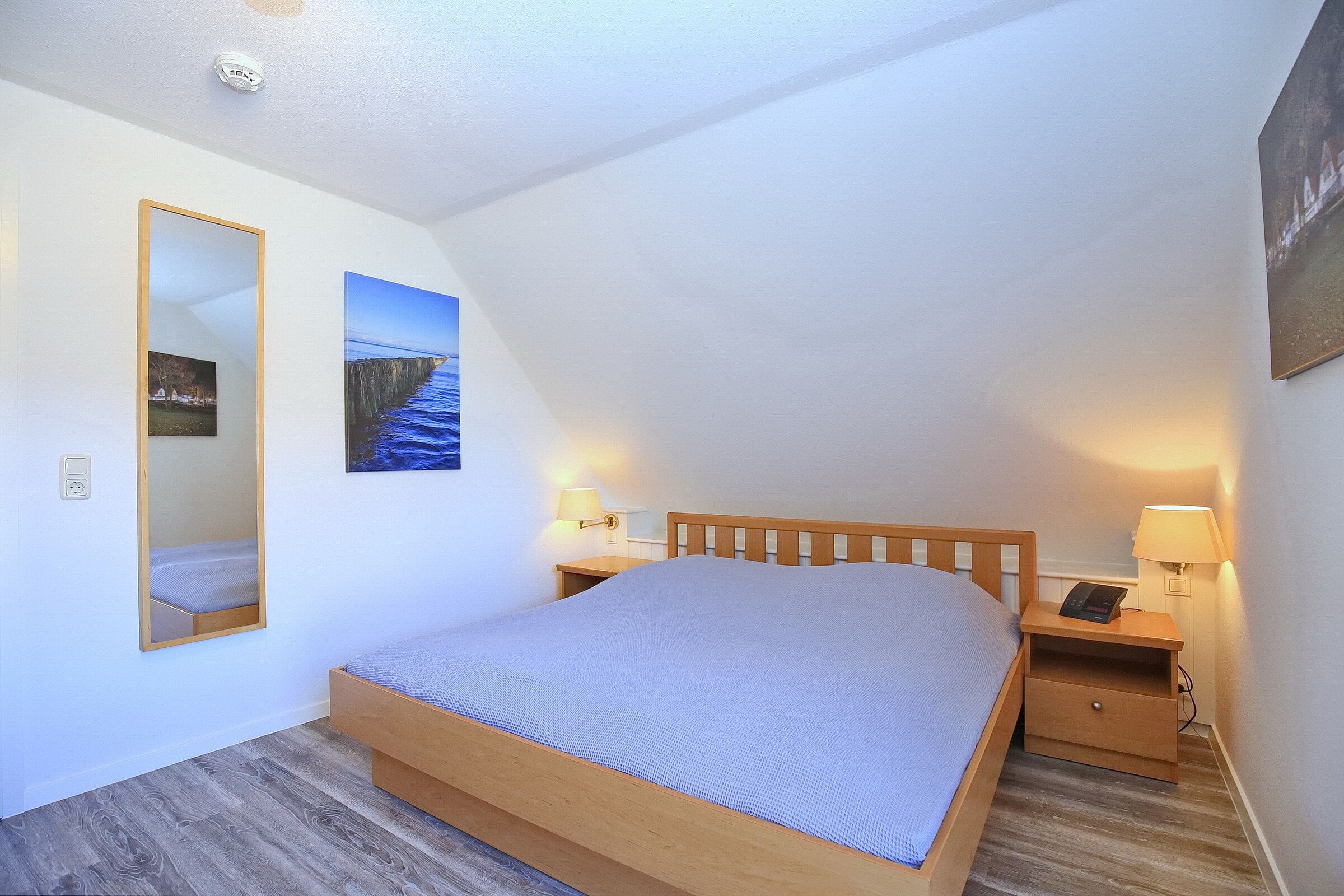 Ein Schlafzimmer mit einem hölzernen Bett, einem Nachttisch mit Lampe, einem wandmontierten Spiegel und einem gerahmten Bild.