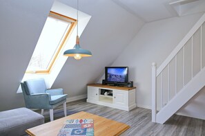 Una habitación estilo loft con techo inclinado, un televisor sobre un mueble de madera, un sillón azul, una mesa de centro de madera y una escalera.