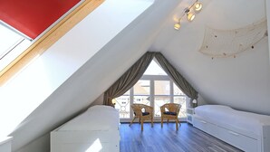 A high-ceilinged room with a sloped ceiling, a large window with curtains, a bed, two wicker chairs, and a white bedside table.