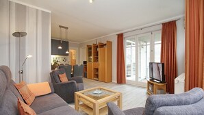 Una sala de estar moderna con un sofá gris, sillones, una mesa de centro y un televisor. Se aprecia una cocina con armarios y una mesa de comedor al fondo.