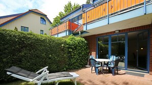 Eine Terrasse mit zwei Liegestühlen und einem Tisch, einer Hecke und einem Gebäude mit Balkon.