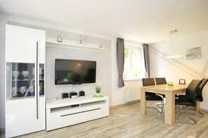 Ein modernes Wohnzimmer mit Flachbildfernseher, einem Esstisch mit Stühlen und einer weißen Unterhaltungsecke mit Glastüren, in denen Weingläser präsentiert werden.