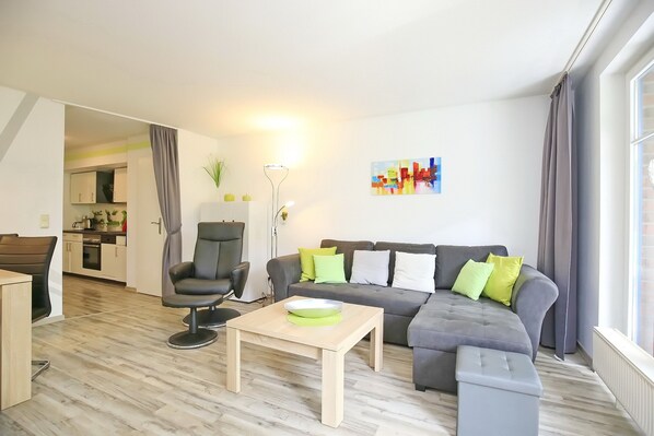 Ein modernes Wohnzimmer mit einem grauen Ecksofa, einem schwarzen Bürostuhl, einem Holztisch und einem grauen Hocker. Im Hintergrund ist eine Küche zu sehen.