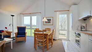 A modern kitchen with white cabinets, a dining area with a wooden table and chairs, and large windows offering a view of the outdoors. - Die Ferienwohnung 21 in der Anlage Godewind ist Eine 3-zimmerwohnung für bis zu (Ostseebad Boltenhagen)