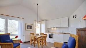 A modern living room with wicker furniture, a dining table, and a kitchen area with white cabinets. - Die Ferienwohnung 21 in der Anlage Godewind ist Eine 3-zimmerwohnung für bis zu (Ostseebad Boltenhagen)