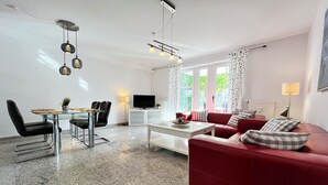 A modern living room with a red sofa, a glass coffee table, and a TV. There is a dining area with a table and chairs, and a kitchen area with a microwave and a plant. - Strandvilla Niklot Wohnung 01 (Ostseebad Boltenhagen)