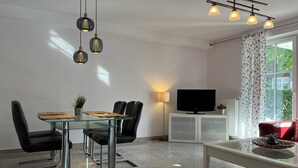 A modern living room with a glass dining table, black leather chairs, a flat-screen TV, and a large window with curtains. - Strandvilla Niklot Wohnung 01 (Ostseebad Boltenhagen)