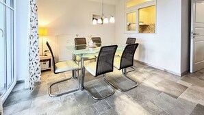 A dining area with a glass table, four chairs, a lamp, and a view of the kitchen through a window.