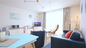 Moderni olohuone, jossa on sininen sohva, ruokapöytä ja televisio. Keittiöalueella on kaappeja ja jääkaappi.
