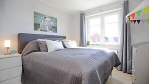 A neatly made bed with a gray quilt, a bedside table with a lamp, a painting on the wall, and a window with curtains. - Villa Bernstein Wohnung 06 (Ostseebad Boltenhagen)