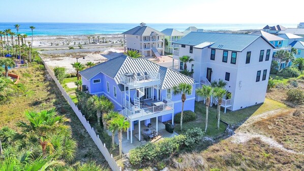 A large, multi-story beachfront property with a prominent balcony area, a covered patio, and a view of the ocean.