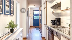 A narrow hallway with a kitchen on one side, featuring a stove, coffee maker, and cabinets. A clock and framed pictures are visible on the wall.