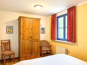 Ein Schlafzimmer mit einem Bett, einem Holzschrank, einem Sessel, einem Fenster mit Vorhängen und zwei an der Wand hängenden Bildern.