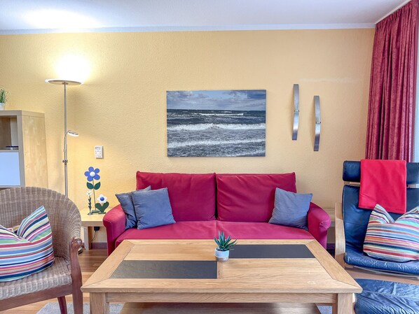 Ein Wohnzimmer mit einem roten Sofa, einem Holztisch, einem Korbstuhl und einem blauen Sessel.