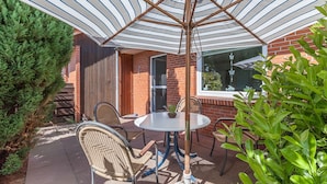 Une terrasse avec une table ronde et des chaises, un parasol à rayures et, au fond, un bâtiment en briques.