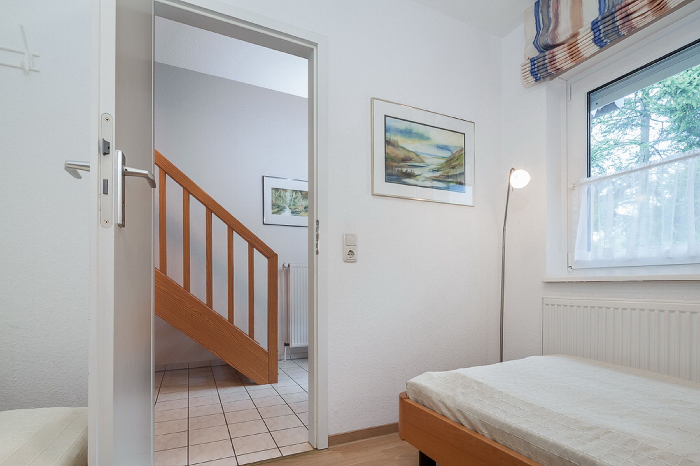 A bedroom with a bed, a window with a view, a door leading to a staircase, and a painting on the wall.