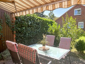 Eine überdachte Terrasse mit einem Glastisch und gestreiften Kissen, umgeben von Grünpflanzen und einem Backsteinhaus im Hintergrund.