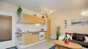 Una cocina moderna con armarios de madera, un microondas y una cafetera. Hay una zona de comedor con una mesa y sillas, y una sala de estar con un sofá y un cuadro en la pared.