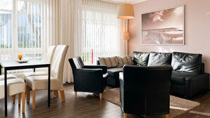 Een moderne woonkamer met een zwarte leren bank, witte stoelen, een eettafel met een kaars en een groot raam met gordijnen.