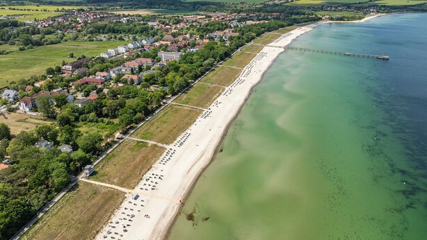 Luchtbeeld van een kustplaatsje met een lang zandstrand, een pier die in het water uitsteekt, en een mix van woon- en winkelgebouwen.