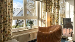 Une pièce avec un fauteuil en osier, une fenêtre ornée de rideaux à motifs floraux, une plante en pot et une vue sur les bâtiments à l’extérieur.