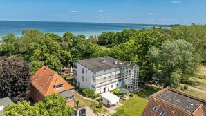 A multi-story building with a flat roof, surrounded by lush greenery and a view of the sea. - Likedeeler II Wohnung 2 (Ostseebad Boltenhagen)