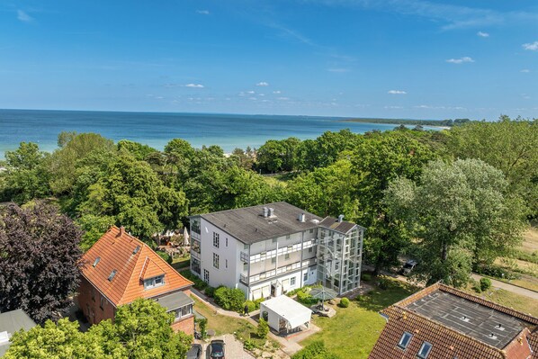 A multi-story building with a flat roof, surrounded by lush greenery and a view of the sea.