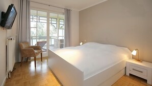 Un dormitorio con una cama grande, una silla, una mesita de noche con lámpara, un televisor montado en la pared y una ventana con cortinas.