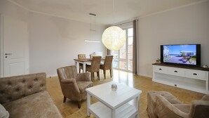 Una sala moderna con un sofá, sillones, una mesa de comedor con sillas, un televisor sobre un mueble bajo y un ventanal con cortinas.