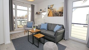 Una sala de estar moderna con un sofá gris, una mesa de centro de madera y una alfombra gris. Hay un ventanal con cortinas, un radiador y un estante en la pared con objetos decorativos.