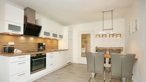 Una cocina moderna con armarios blancos, horno empotrado, cafetera y zona de comedor con mesa de madera y sillas grises.