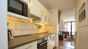 Eine kompakte Küche mit weißen Schränken, einer Mikrowelle, einem Backofen und einem Spülbecken. Durch einen offenen Durchgang ist eine Essecke mit einer Couch und einem Sessel zu sehen.