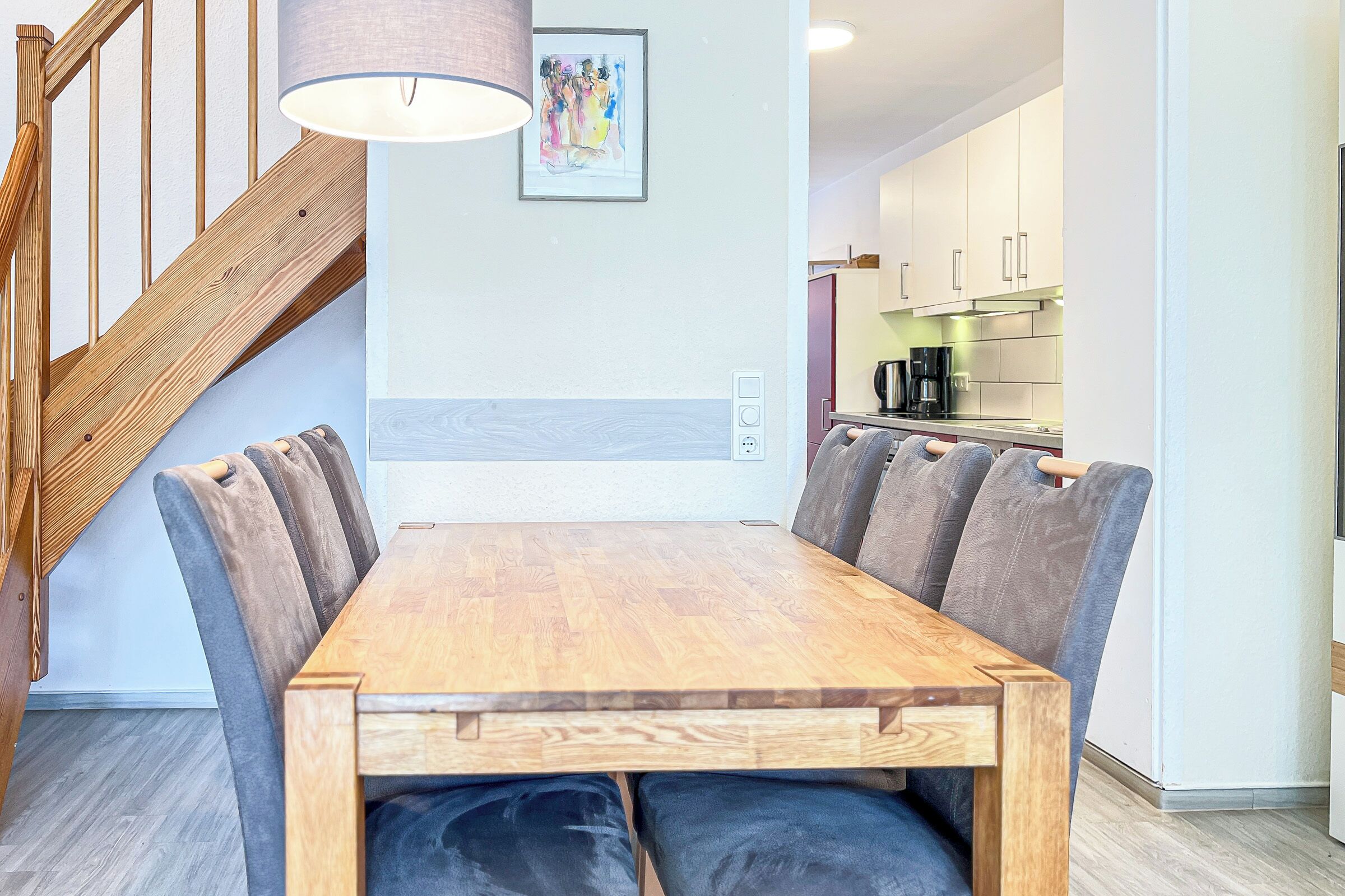 Ein Essbereich mit Holztisch und gepolsterten Stühlen, eine Treppe mit Holzhandläufen und eine Küche im Hintergrund.