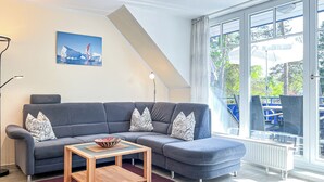 Ein modernes Wohnzimmer mit einem grauen Ecksofa, einem Holztisch und einem großen Fenster mit Blick auf Gartenmöbel.